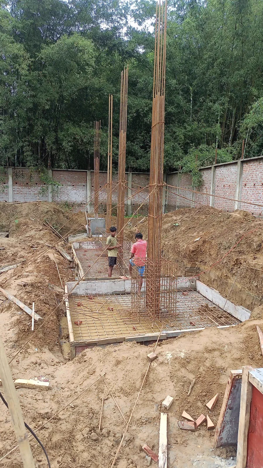 Excavated footing pit with a reinforcement mat tied and a column cage rising from it, bamboo grove behind