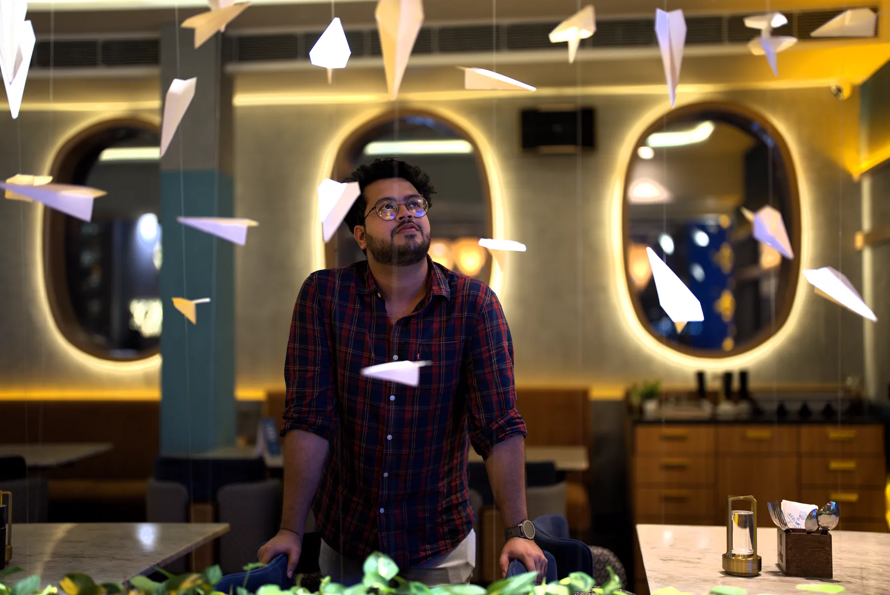 Ritiman Mukherjee at Aerosky, looking up at the paper planes suspended beneath the ceiling, the backlit porthole windows behind him